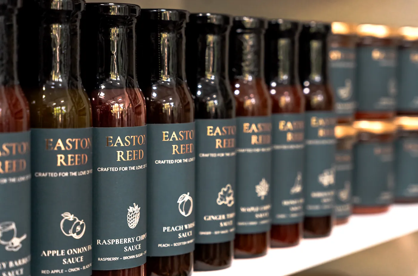 Row of Easton Reed sauce bottles on a shelf: Apple Onion Fig, Raspberry Chipotle, Ginger Teriyaki, Peach Whiskey and Maple Chipotle